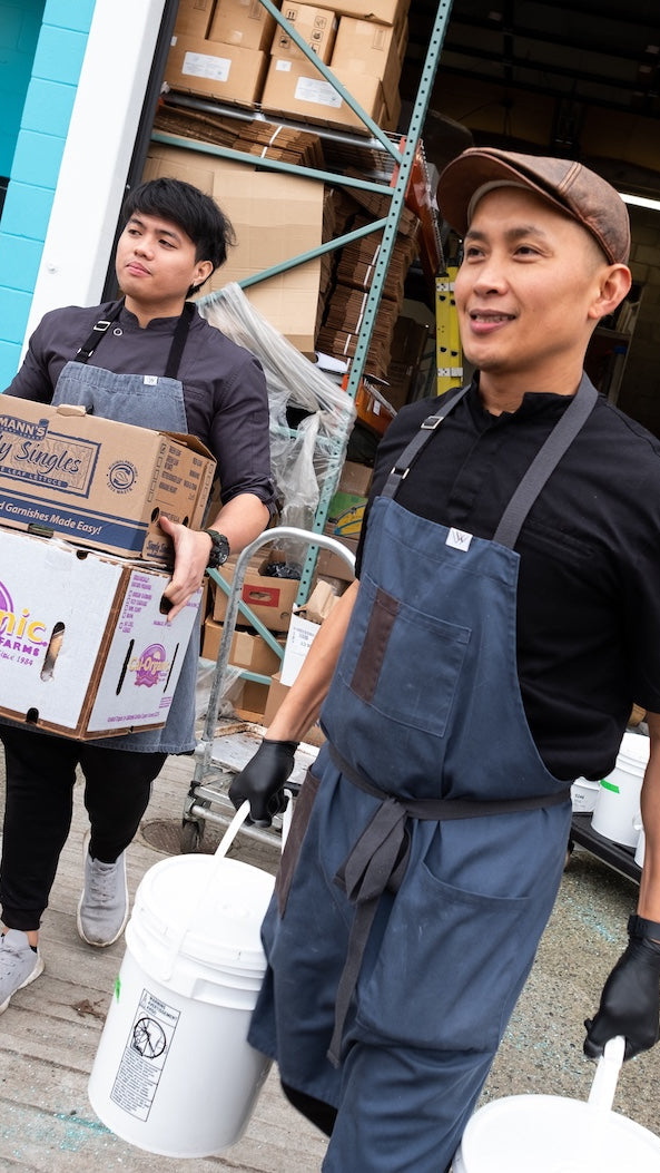 Two workers in a warehouse setting, one holding boxes and the other with a bucket.