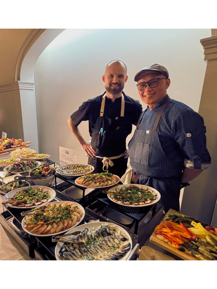 Two chefs standing behind a counter with various dishes in a kitchen setting.